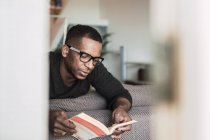 Сосредоточенный афроамериканец в очках читает книгу, сидя дома на диване — стоковое фото