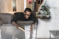 Сосредоточенный афроамериканец в очках читает книгу, отдыхая дома на диване — стоковое фото