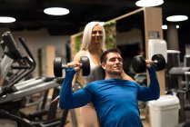 Femme aidant l'homme faisant de l'exercice avec des haltères dans la salle de gym — Photo de stock