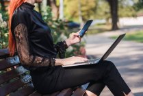 Femme utilisant ordinateur portable et smartphone sur banc dans le parc — Photo de stock