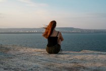 Vue arrière de jeune femme assise sur un rivage en béton et admirant une belle vue sur la mer calme par temps venteux en bulgarie, balkans — Photo de stock