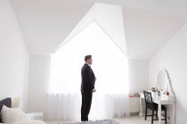 Seitenansicht eines jungen gutaussehenden Mannes, der in einem weißen Schlafzimmer steht und ein schwarzes formales Kostüm anzieht — Stockfoto