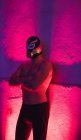 Homme musclé sans chemise portant un masque de combattant et debout avec les bras croisés en lumière rouge — Photo de stock