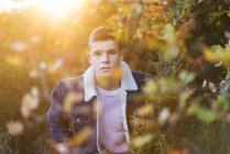Portrait of teenage boy in denim jacket standing in sunny bushes — Stock Photo