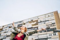 Redhead young woman with arms outstretched standing against residential building — Stock Photo
