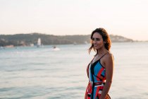 Conteúdo mulher em vestido colorido andando no calçadão calçadão ao pôr do sol contra a paisagem marinha — Fotografia de Stock