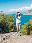Young woman in shorts using mobile phone and taking selfie standing on cliff with trees on background of blue seascape — Stock Photo