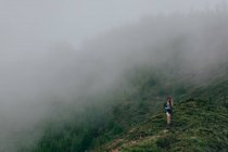 Женщина идет по высокому крутому холму, покрытому зеленой травой с густым туманом. — стоковое фото