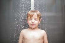 Rubio chico con los ojos cerrados de pie bajo el agua en la ducha - foto de stock