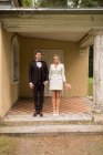 Красивий модний чоловік і жінка у весільних костюмах тримаються за руки і стоять на ганку старого будинку — стокове фото