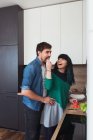 Vista lateral de la joven emocionada dando tomate cortado a novio alegre mientras se cocina en la cocina con estilo juntos - foto de stock