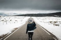 Rückansicht eines jungen Menschen in stylischem Outfit, der an bewölkten Wintertagen mitten auf einer asphaltierten Straße in einer wunderschönen Landschaft spaziert — Stockfoto