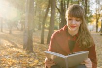 Jolie jeune femme en manteau rouge tenant livre ouvert et assis sur le siège dans la forêt d'automne — Photo de stock