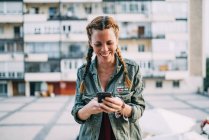 Усміхнена руда дівчина з косами, використовуючи мобільний телефон проти житлового будинку — стокове фото