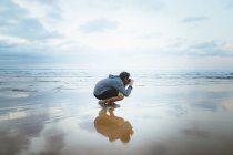 Vista laterale di anonimo giovane ragazzo seduto sulla sabbia bagnata vicino al mare incredibile e scattare foto nella giornata nuvolosa a Zarautz, Spagna — Foto stock