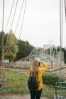 Back view of blond woman with camera taking photo of desolated amusement park with attractions — Stock Photo