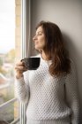 Mulher adulta bonita em camisola de malha sorrindo e olhando para longe enquanto segurando xícara de bebida quente fresca e inclinando-se na parede — Fotografia de Stock
