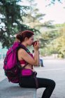 Junge Frau in Sportbekleidung mit rosa Rucksack sitzt auf Bank im Park und trinkt heißen Tee aus Metallbecher — Stockfoto