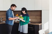 Jovem e mulher em trajes casuais sorrindo e segurando tigela com alface fresca enquanto cozinham na cozinha moderna juntos — Fotografia de Stock