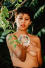 Giovane donna bruna in topless che copre il seno e tiene la palla di vetro trasparente in boschi verdi — Foto stock