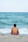 Вид сзади мужчины в шортах, сидящего на песчаном берегу возле великолепного моря, машущего в Тыленово, Болгария — стоковое фото