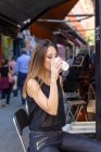 Attraktive Frau in stylischem Outfit hält die Augen geschlossen und genießt frisches Heißgetränk, während sie auf dem Stuhl auf dem Markt sitzt — Stockfoto