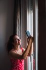 Jovem mulher tomando selfie com telefone celular ao lado da janela em casa — Fotografia de Stock