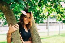 Mujer alegre y elegante de pie bajo el árbol abrazando el tronco y sonriendo a la cámara en el parque - foto de stock