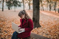 Young lady in red coat using device and sitting on seat in autumn forest — Stock Photo