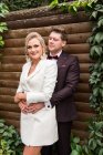 Eleganti sposi adulti che si abbracciano in piedi in giacca e cravatta contro la parete di legno e sorridono alla macchina fotografica — Foto stock