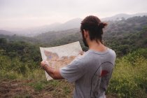 Vista posteriore di ragazzo anonimo in abito casual in piedi nella natura incredibile e guardando la mappa nella giornata nebbiosa — Foto stock