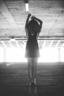 Vue arrière de la mince femme bouclée portant une robe noire avec des baskets debout sur un parking souterrain à la lumière du soleil — Photo de stock