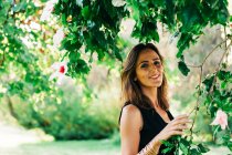 Cheerful stylish woman standing under tree and smiling at camera in park — Stock Photo