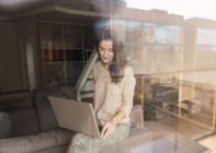 Досить доросла жінка в елегантному вбранні, переглядаючи сучасний ноутбук і дивлячись на камеру, сидячи в затишній кімнаті за величезним склом вікна — стокове фото