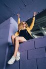 Élégante femme blonde bouclée en baskets et veste jaune assise sur le mur violet et riant — Photo de stock