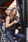 Atractiva hembra con taza de bebida caliente y teléfono inteligente moderno sonriendo y mirando a la cámara mientras está sentada cerca de la cafetería - foto de stock