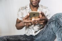 Крупный план блестящего современного мобильного телефона в руках черного человека, сидящего у белой стены — стоковое фото