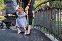 Женщина в туфлях на высоких каблуках учит милую малышку ходить по небольшому мосту в парке — стоковое фото