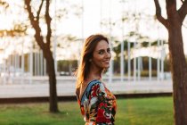 Lächelnde stylische Brünette im Kleid, die im Stadtpark steht und in die Kamera schaut — Stockfoto