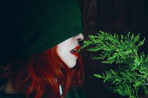 Joven mujer pelirroja con labios rojos y sombrero verde mordiendo ramita de abeto - foto de stock