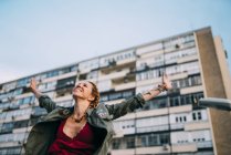 Redhead young woman with arms outstretched standing against residential building — Stock Photo