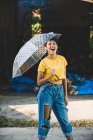 Charming young woman in stylish outfit laughing and holding transparent umbrella while standing on street on sunny day — Stock Photo