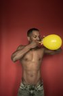 Muskulöse hemdlose schwarze Mann in Jeans weht leuchtend gelben Ballon auf rotem Hintergrund — Stockfoto