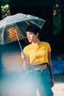 Adorável jovem mulher em roupa elegante segurando guarda-chuva e olhando para longe, enquanto de pé na rua durante a chuva no dia ensolarado — Fotografia de Stock