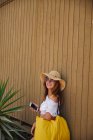 Весела жінка з коричневим волоссям і сонцезахисними окулярами в білому верхньому і солом'яному капелюсі тримає жовтий мішок і пристрій на дерев'яному фоні стіни і зеленому кущі — стокове фото