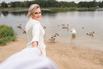 Adult beautiful and elegant bride holding crop hand of groom and smiling at camera while standing on lake coast with swans — Stock Photo