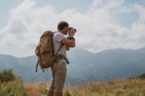 Vista trasera del hombre con la mochila usando la cámara profesional para hacer fotos del pintoresco campo en Bulgaria, Balcanes - foto de stock