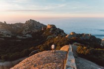 Vista trasera del hombre en traje casual caminando en hermosas montañas hacia el mar tranquilo durante el increíble amanecer en Barcelona, España - foto de stock