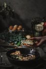 Vista de la mano con sabrosa tortilla en la sartén cerca de platos con rebanadas, tomates, frutas, nueces y hojas sobre tabla de madera - foto de stock