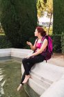 Junge Frau mit Rucksack sitzt mit Kompass in der Hand auf Brüstung am See — Stockfoto
